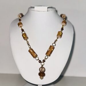 Tan paper based beads with dashes, moons and stars, connected with gold fixings, alternating with brown and copper toned seed beads and an artist made charm of stone and glass bars, on a white body form with a neutral background