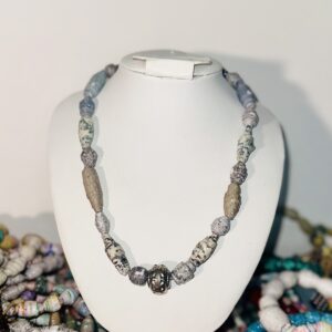 A white necklace form set in the midst of a variety of bracelets and displaying a gray and silver paper-bead necklace with a metal charm bead.