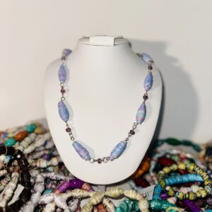 A white necklace form set amidst a collection of bracelets and displaying a paper-bead necklace of lavender and blue with glass seed beads of purple.