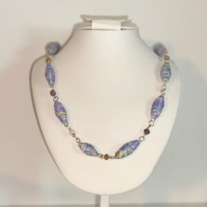Blue beads with tiny flowers with glass beads and silver fixings on a white necklace stand with a beach background