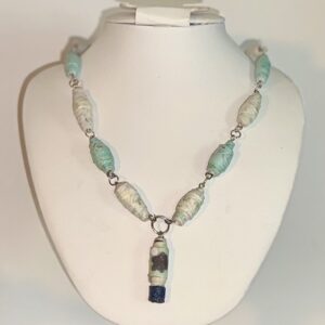 Necklace of various blue and blue flower paper beads with a large artist made charm and silver fixings on a white form with a beach background